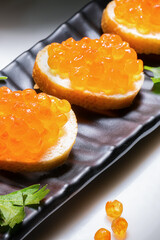 Red caviar on butter buns on black rectangular plate of parsley is illuminated by very bright white light. Caviar on butter rolls is illuminated by light.