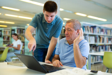 Young guy helping older man in laptop interface in public library