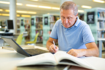 Focused casual mature male writing in notepad and reading books in the library. High quality photo