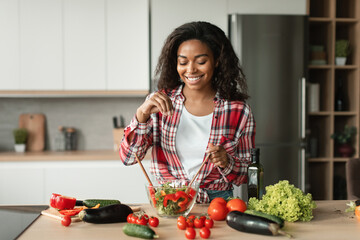Homemade food and health care. Cheerful black young woman chef in casual salts salad from organic vegetables