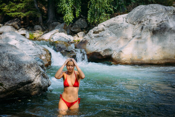 Beautiful young woman wearing red bikini coming out stormy mountain river. Happy female brunette with sexy body on picturesque natural background.