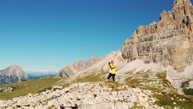 Blonde woman takes pictures of mountain tops standing on high rocky hill. Female tourist raises hands up triumphantly near Tre Cime di Lavaredo aerial view
