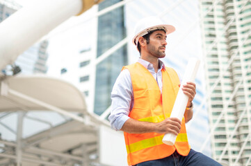 Young caucasian man holding a big paper, a guy wearing a light blue shirt and jeans with an orange vest and white helmet for security in a construction area.