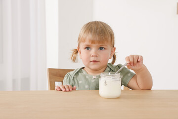 Cute little child eating tasty yogurt with spoon at wooden table indoors