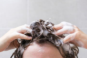 A girl washes her hair with shampoo on gray tiles background, front view. - obrazy, fototapety, plakaty
