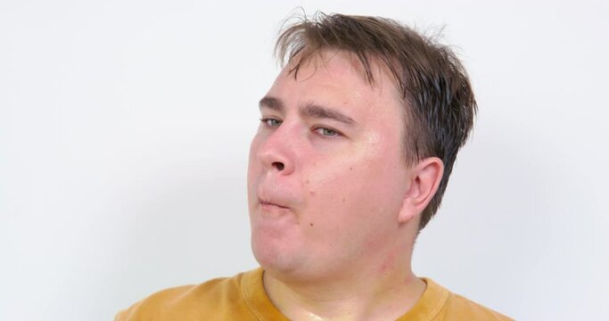 Mature plump man cools rubbing face with ice cube sitting on comfortable sofa at home on hot summer day. After rubbing face guy puts ice cube into mouth