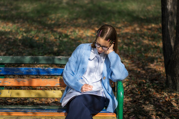 A teenager on a bench is doing homework on a sunny autumn day.