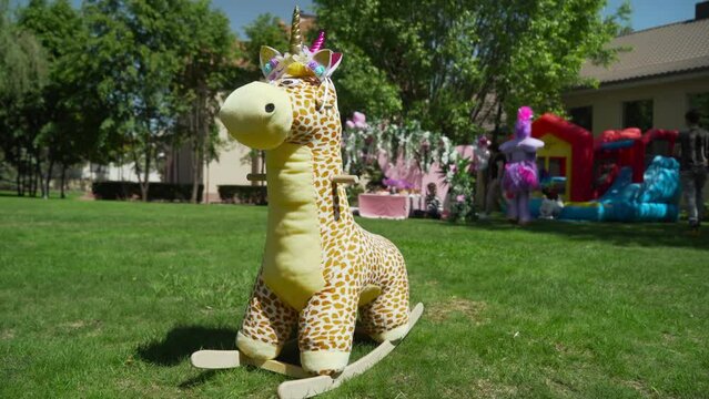 A large stuffed giraffe toy is lying on the lawn. A children's party is taking place in the background.