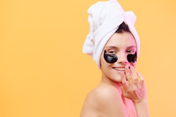 Horizontal photo, a woman with perfect skin on an orange background in a towel on her head and body with black patches on her face looks away and plays with her hands