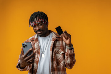 Obraz premium Smiling young african man on yellow background using mobile phone and holding credit card.