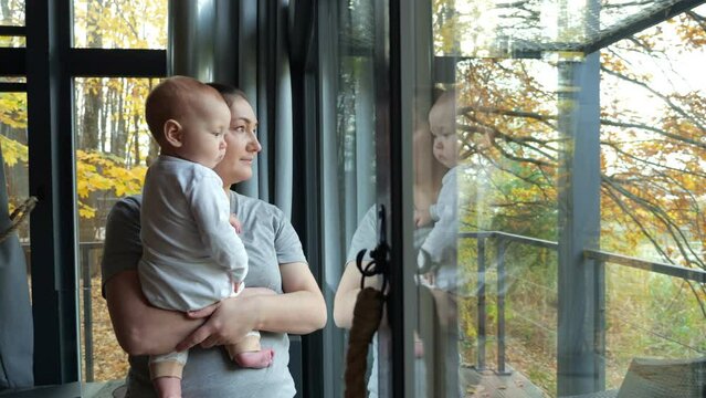 Young mother stands near window holding baby in arms. Child looks with interest stretching out hand to window. Woman smiles and speaks gently