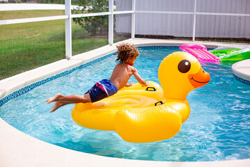 Young diverse little boy jumping onto a large inflatable pool toy in a backyard swimming pool on a...