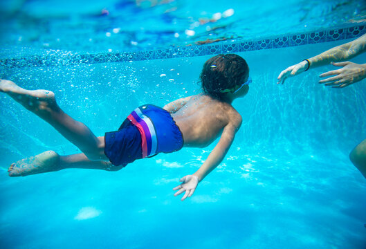 Young diverse boy swimming underwater in a swimming pool. Learning to swim with the help of his parent. Photo below the water showing a child practicing diving under the water