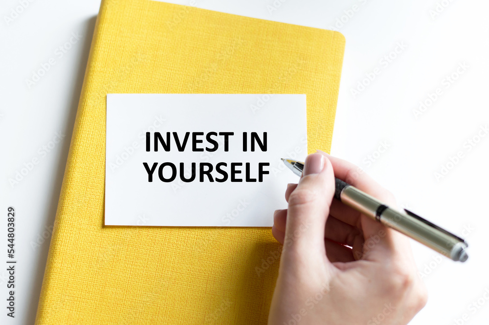Poster invest in yourself text on a card on a yellow notebook next to the hand of the bang writes the text