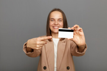 Photo of satisfied brown-haired woman wearing jacket pointing at plastic credit card, recommend bank or shopping discounts, looking at camera with toothy smile, posing isolated over gray background.