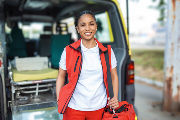 Young woman , a paramedic, standing at the rear of an ambulance, by the open doors. She is looking at the camera with a confident expression, smiling, carrying a medical trauma bag on her shoulder.