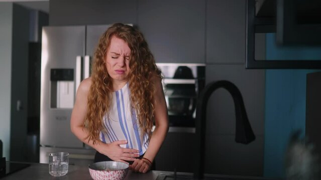 Young woman suffering from severe stomach ache and takes pills while having a meal at kitchen. Female feeling extremely sick prepating meal and takes medicine. Girl suffering from a sharp belly pain.