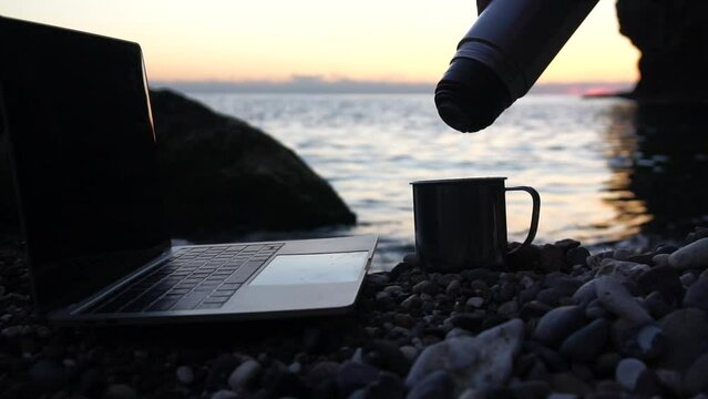 Silhouette of laptop and tea is poured into a cup on the beach over sea sunset. Sparkling sea water on background. Freelancer, blogger working on notebook on the beach.