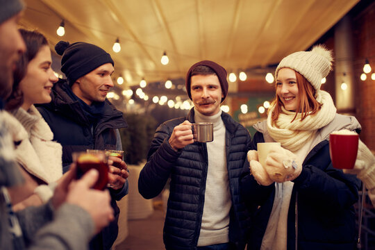 Group of young people spending time together, drinking mulled wine, talking, laughing at winter fair at evening time. Winter holidays, Christmas vibes concept