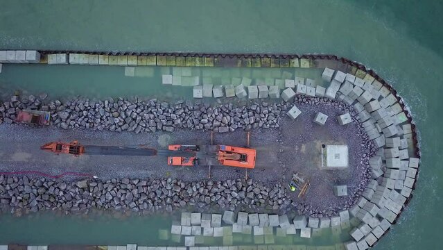 Aerial view of construction site on the Baltic sea shore, strengthening the Baltic Sea coastline, building protective stone pier, heavy duty machinery unloading concrete blocks, birdseye dolly view