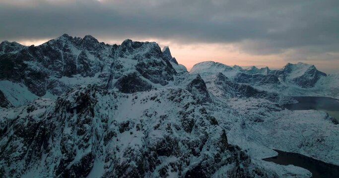 Flying across rugged Lofoten snow covered Svartvatnet head mountains aerial orbiting view with cold sunrise clouds