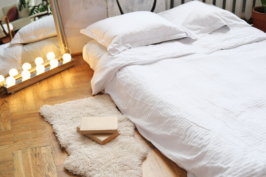 Bedroom interior design. An air mattress with linens on a wooden floor, next to it there is a rug with a stack of books and a stylish mirror with light bulbs