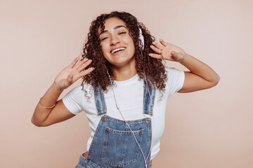 Young cheerful arabic woman girl with curly hair, wears denim overalls, listens mp3 music podcast...