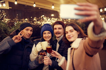 Friendly party. Emotive smiling men and women with mugs of mulled wine having fun, spending time together at winter fair at evening time. Winter holidays, Christmas concept