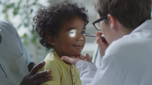 Cute African American boy sitting with father while female doctor checking his eyes with flashlight during checkup in pediatric clinic