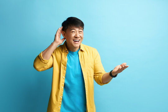 Asian Man Trying to Hear Isolated. Portrait of Confused Guy Keeping Hand Near Ear to Listen Better, Having Hearing Problems, Difficult to Understand. Indoor Studio Shot, Blue Background 