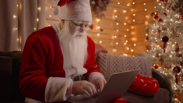 A portrait of a real Santa Claus working on a laptop walking on the couch at home on Christmas Eve. Santa responds to emails, browses the Internet bank and works on a laptop
