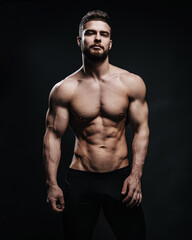 Slim muscular male model at black background. Fitness shirtless guy in black sport pants posing in...
