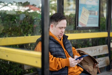 Man sitting on bench at bus stop, using smart phone - obrazy, fototapety, plakaty