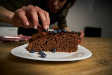 Woman with a passion for cooking decorates a slice of handmade chocolate cake in her home kitchen with blackberries and red currants.