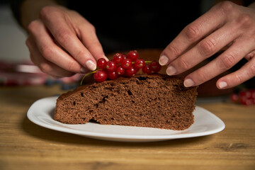 Woman with a passion for cooking decorates a slice of handmade chocolate cake in her home kitchen with blackberries and red currants.