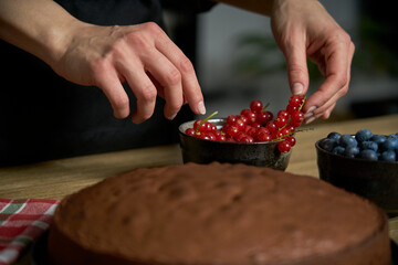 Woman with a passion for cooking decorates a slice of handmade chocolate cake in her home kitchen with blackberries and red currants.