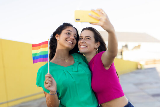 A beautiful lesbian young couple embraces and holds a rainbow flag. Girls taking selfie photo...