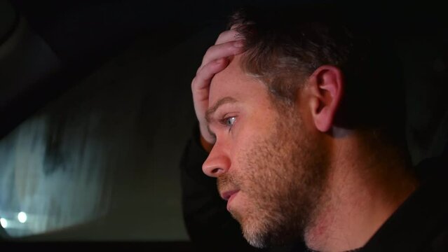 A sad man alone in a car at night with traffic passing on a busy road.