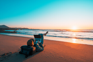 mature latin digital nomad laying on the sand of the beach with a laptop working at sunset over the ocean - obrazy, fototapety, plakaty