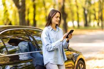 Fototapeta na wymiar Successful smiling attractive woman is using her smart phone while standing near modern car outdoors