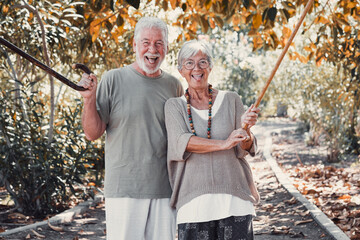 Beautiful senior couple in public park under a shade of large tree while playing with walking sticks sticking their tongues at camera. Carefree caucasian couple enjoying freedom and healthy lifestyle