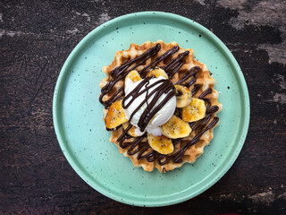 Waffle topped with banana, ice cream,  maple syrup, and chocolate paste served on a round plate. Top view.