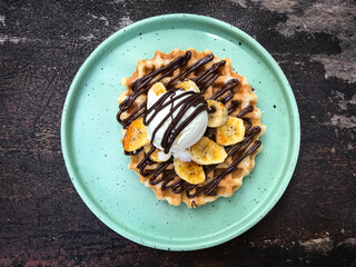 Waffle topped with banana, ice cream,  maple syrup, and chocolate paste served on a round plate. Top view.