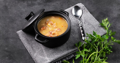 Pea soup with smoked ham in black soucepan on dark background with herb.
