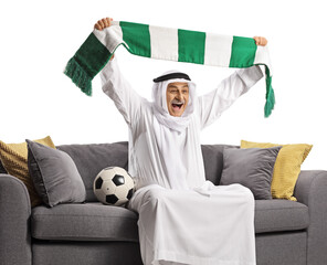 Fototapeta Cheerful arab man cheering with a scarf and sitting on a sofa with a football obraz