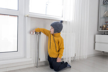 A small child in a yellow sweater and a gray hat is kneeling near a heater with a thermostat.