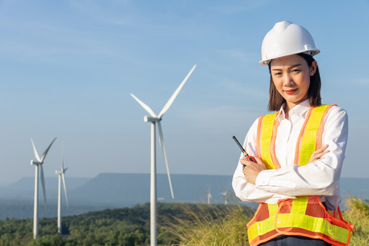 Portrait of beautiful asian woman engineer wearing safety helmet, reflective vest and folded arms and holding walkie talkie with wind turbine propeller background. Environmental friendly concept.