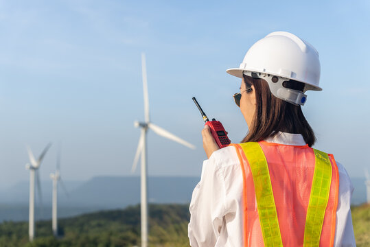 Back view of beautiful asian woman engineer wearing safety helmet and reflective vest talking to her team or someone with radio communication with wind turbine propeller background.