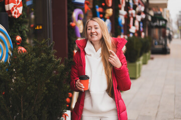 lovely woman in winter clothes walks down the street decorated with New Year's decor and drinks hot coffee