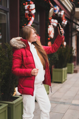 lovely woman in winter clothes walks down the street decorated with New Year's decor and takes selfie on phone